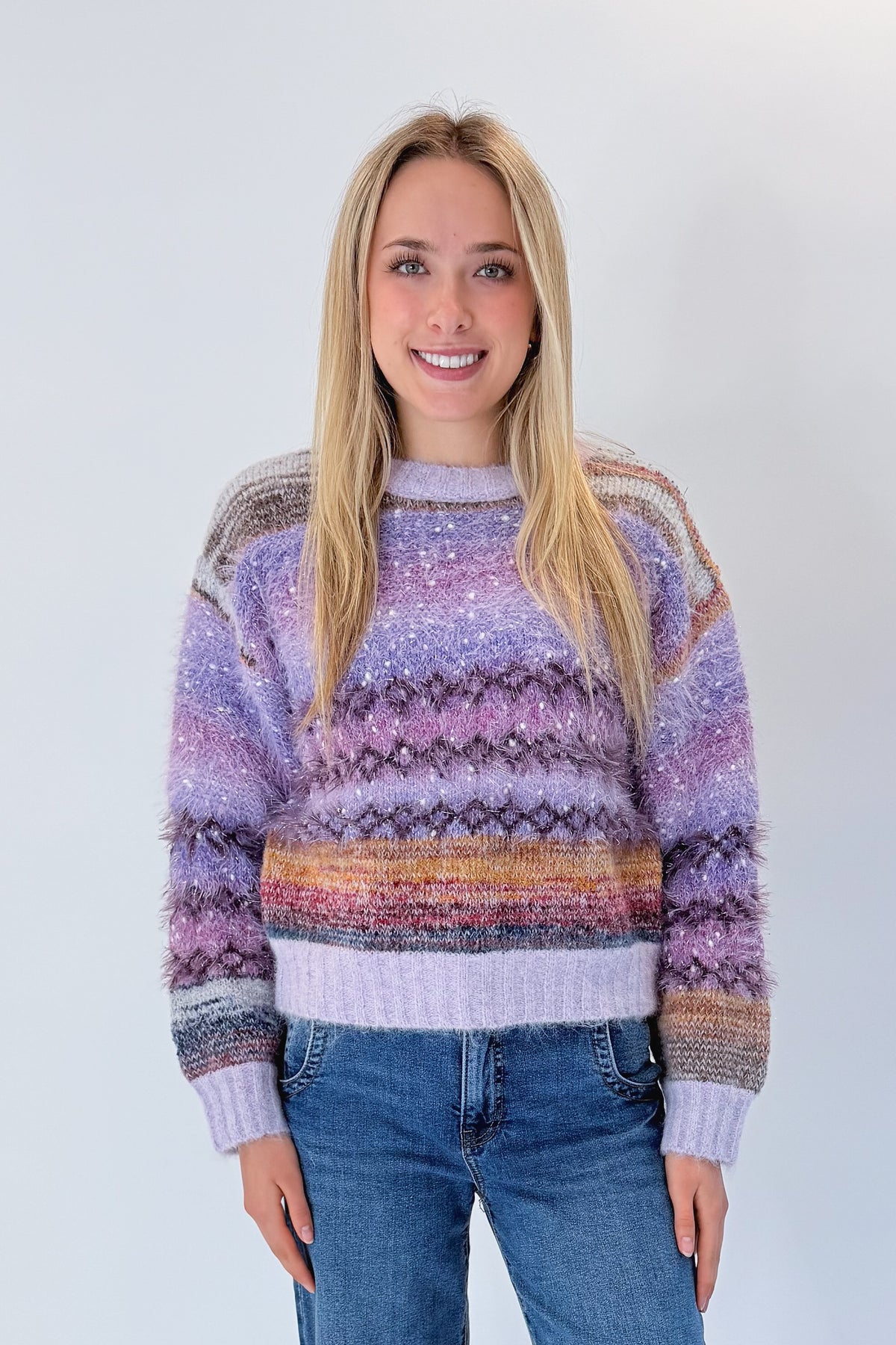 Front view of woman wearing Swagger Boutique’s Purple Printed Eyelash Sweater with jeans against white studio background.