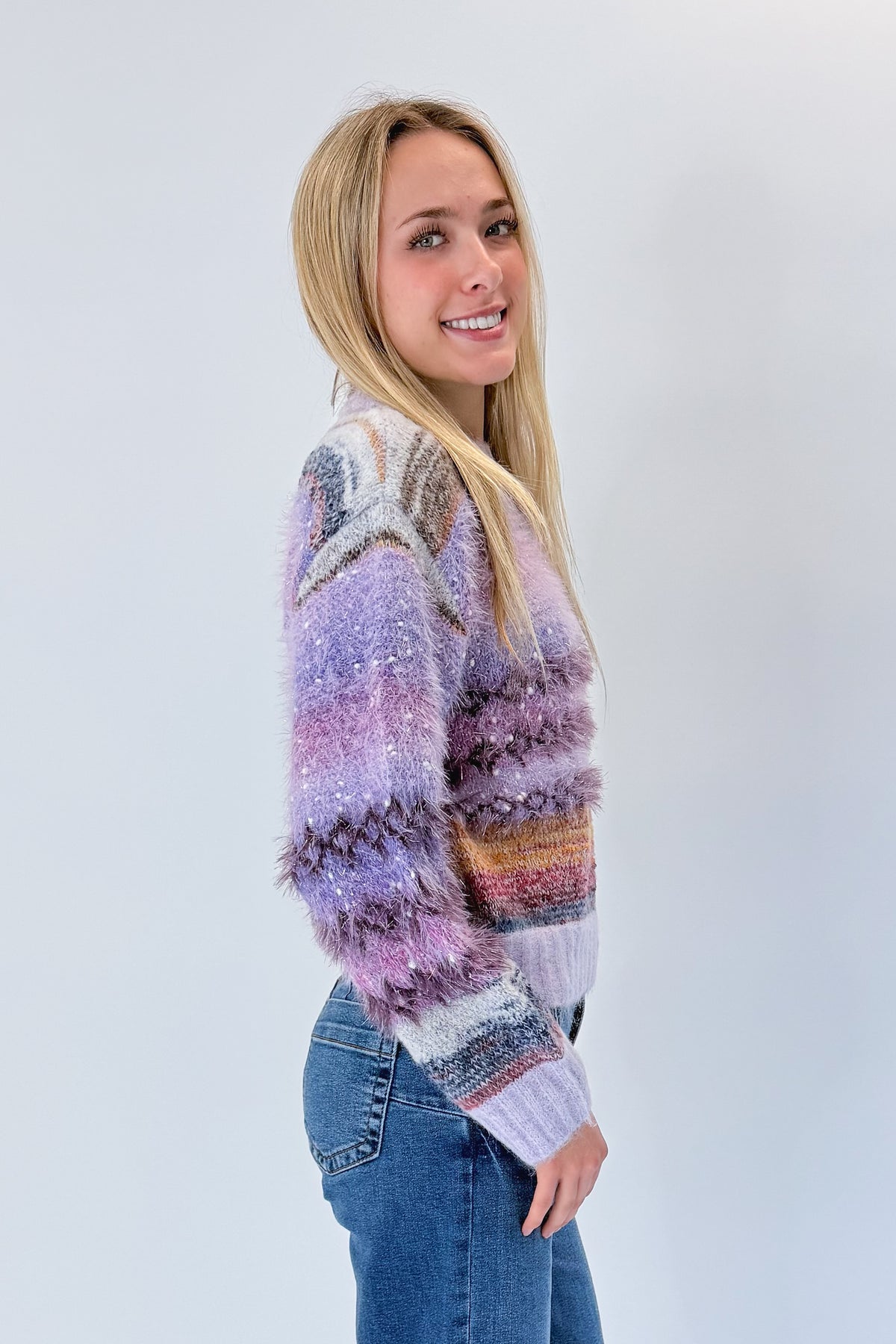 Side view of woman wearing Swagger Boutique’s Purple Printed Eyelash Sweater with jeans against white studio background.