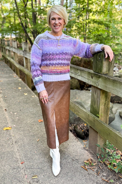 Front view of woman wearing Swagger Boutique’s Purple Printed Eyelash Sweater with Brown Faux Leather Slit Midi Skirt standing outside on a bridge. 