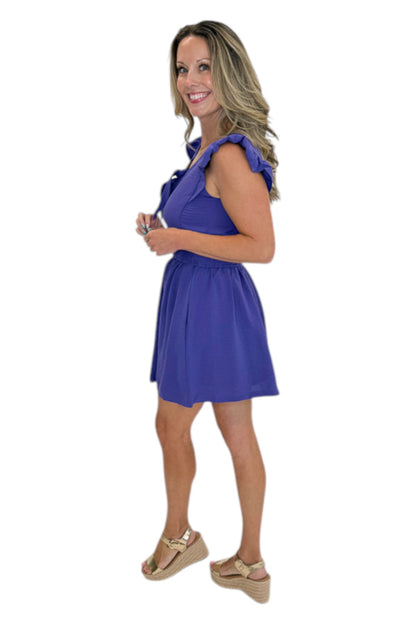 Side view of woman wearing Swagger Boutique’s Purple Tie Detail Smocked Waist Dress with sandals against white studio background.