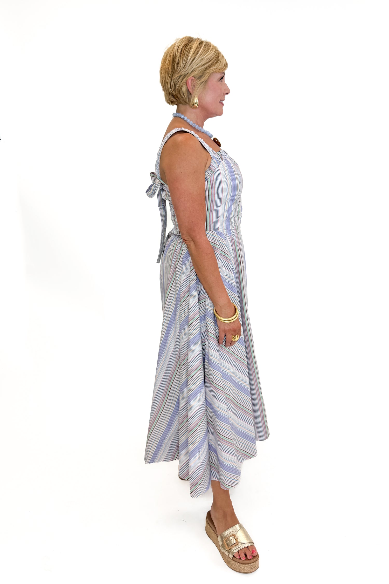 Side view of woman wearing Swagger Boutique’s Purple &amp; Multi Striped Sleeveless Midi Dress with sandals against white studio background.