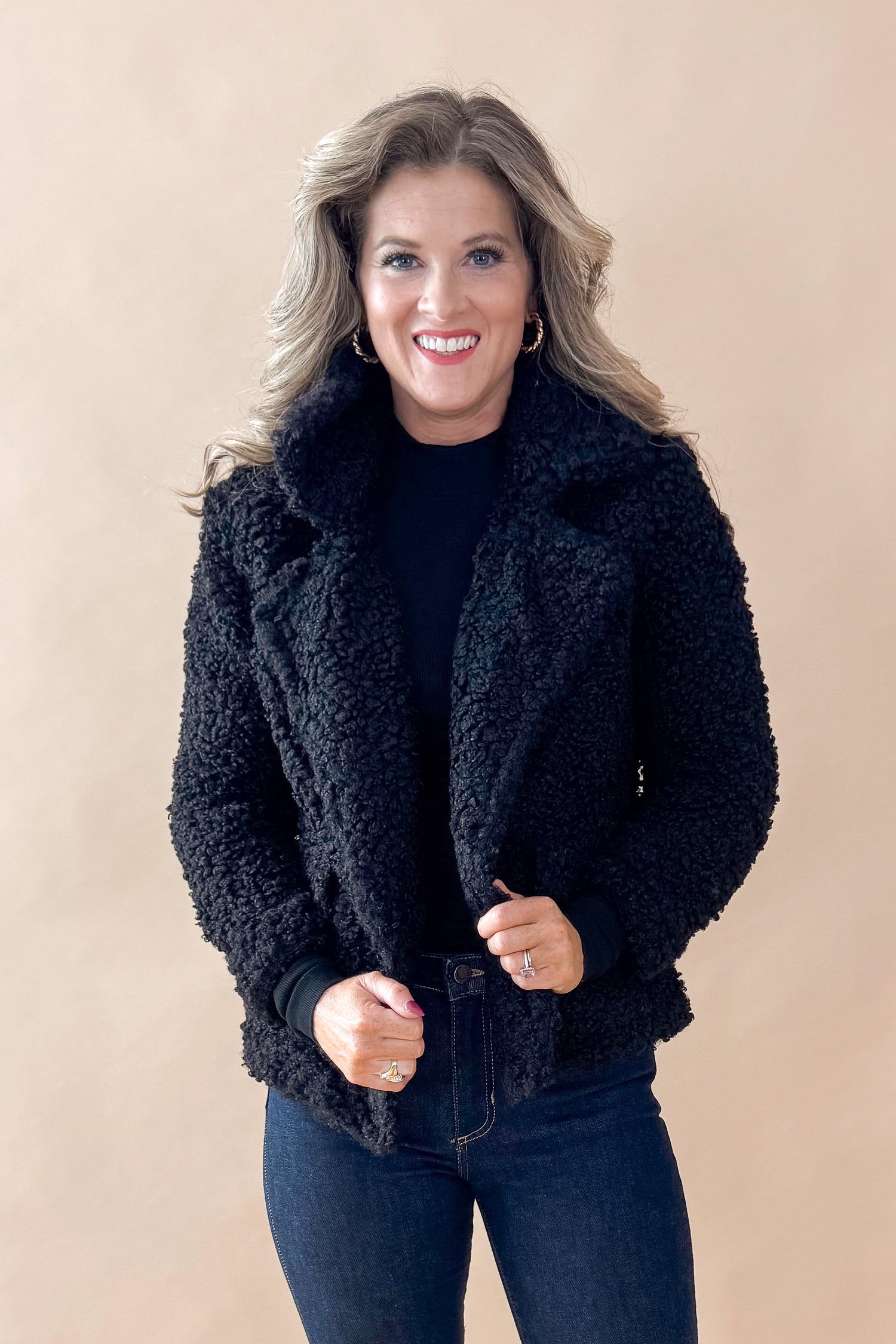 Front view of woman wearing Black Sherpa Coat and jeans against tan studio background.