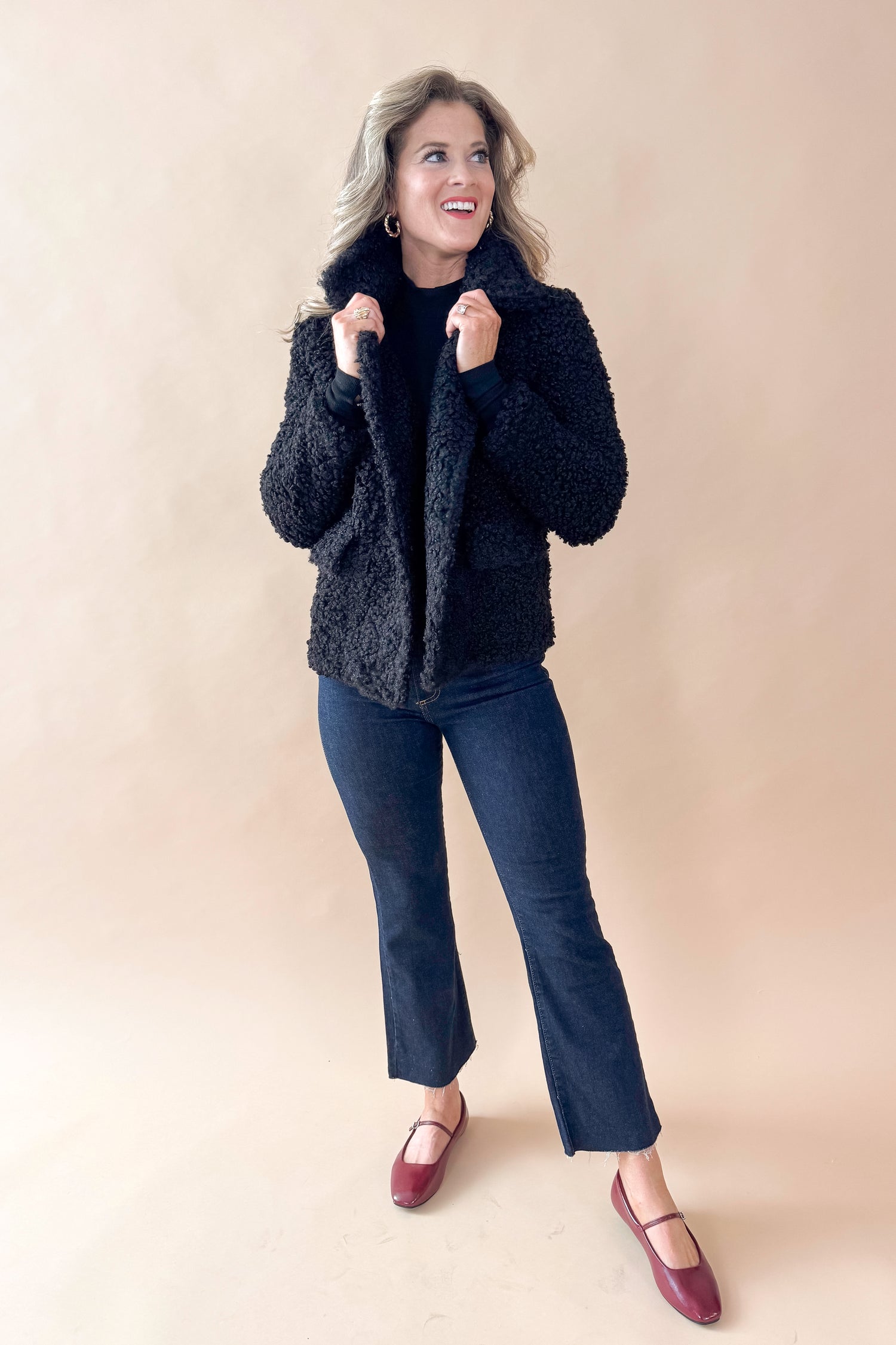 Front view of woman wearing Black Sherpa Coat and jeans against tan studio background.