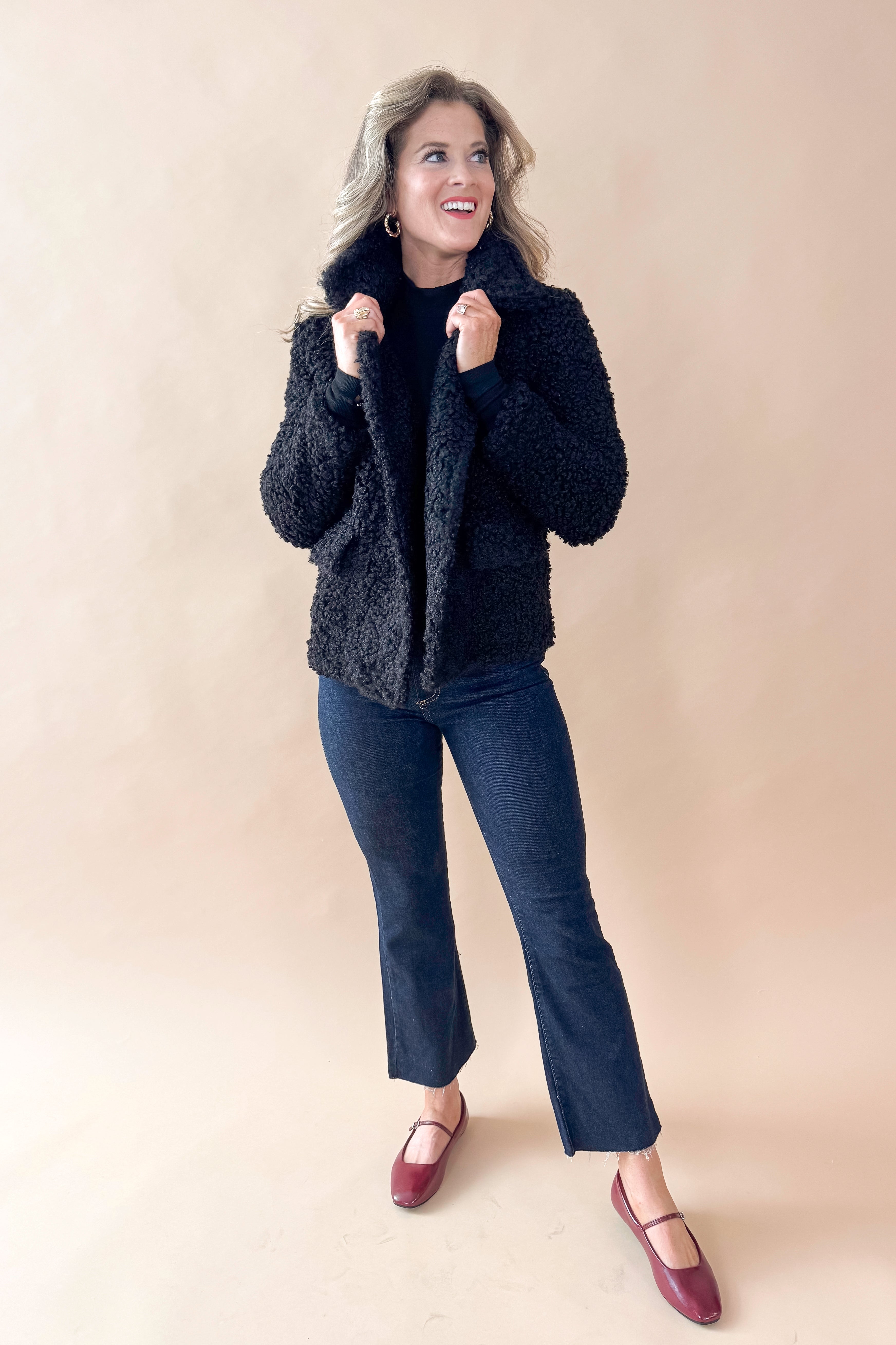 Front view of woman wearing Black Sherpa Coat and jeans against tan studio background.