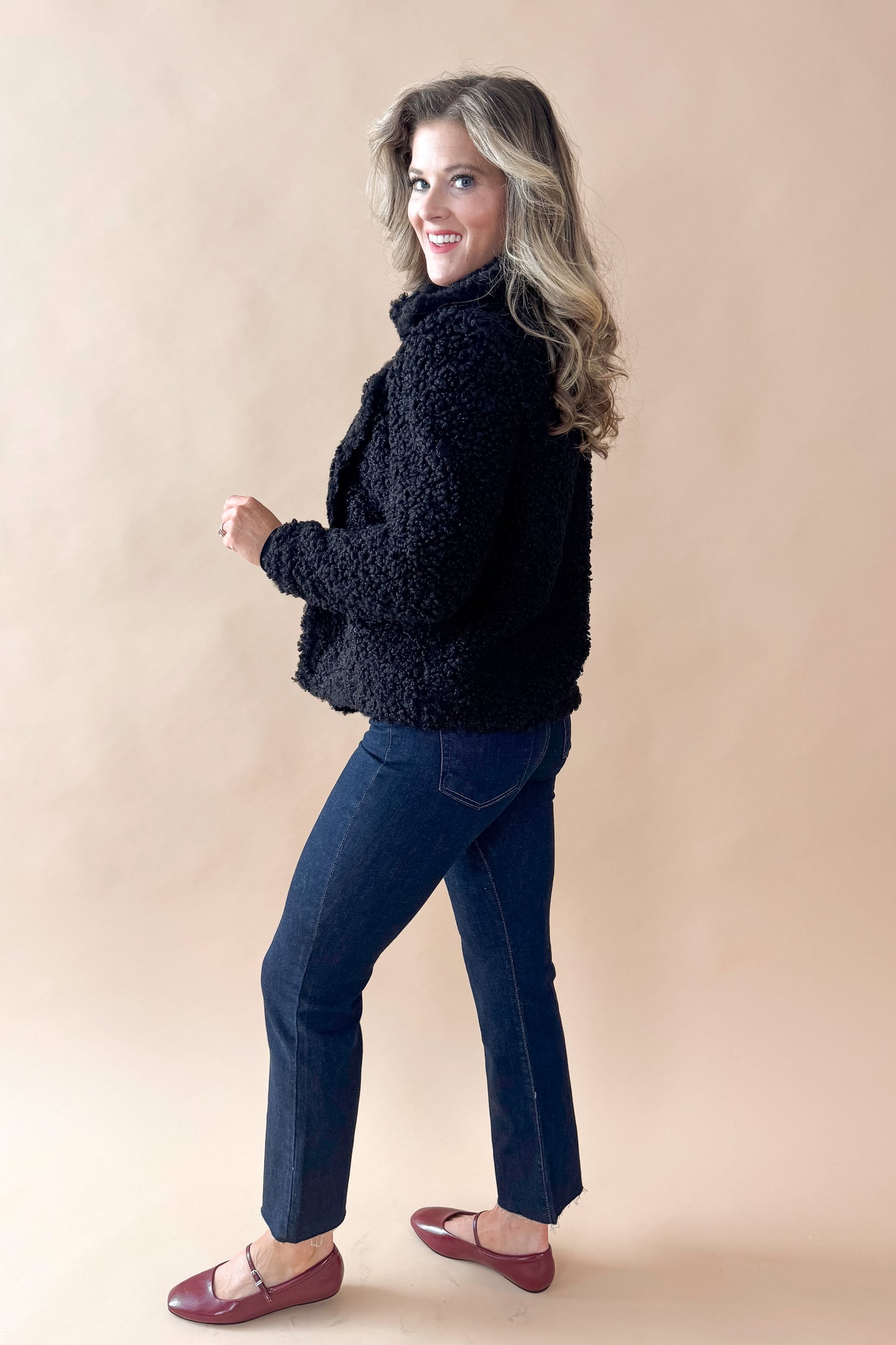 Side view of woman wearing Black Sherpa Coat and jeans against tan studio background.