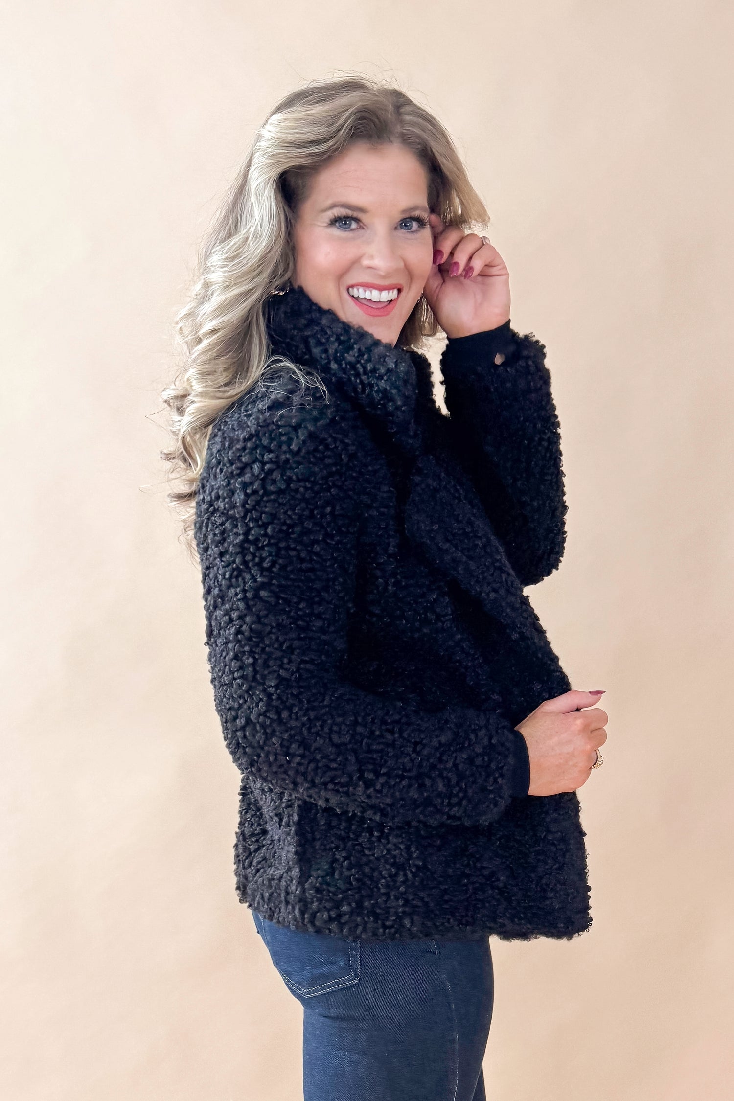 Side view of woman wearing Black Sherpa Coat and jeans against tan studio background.
