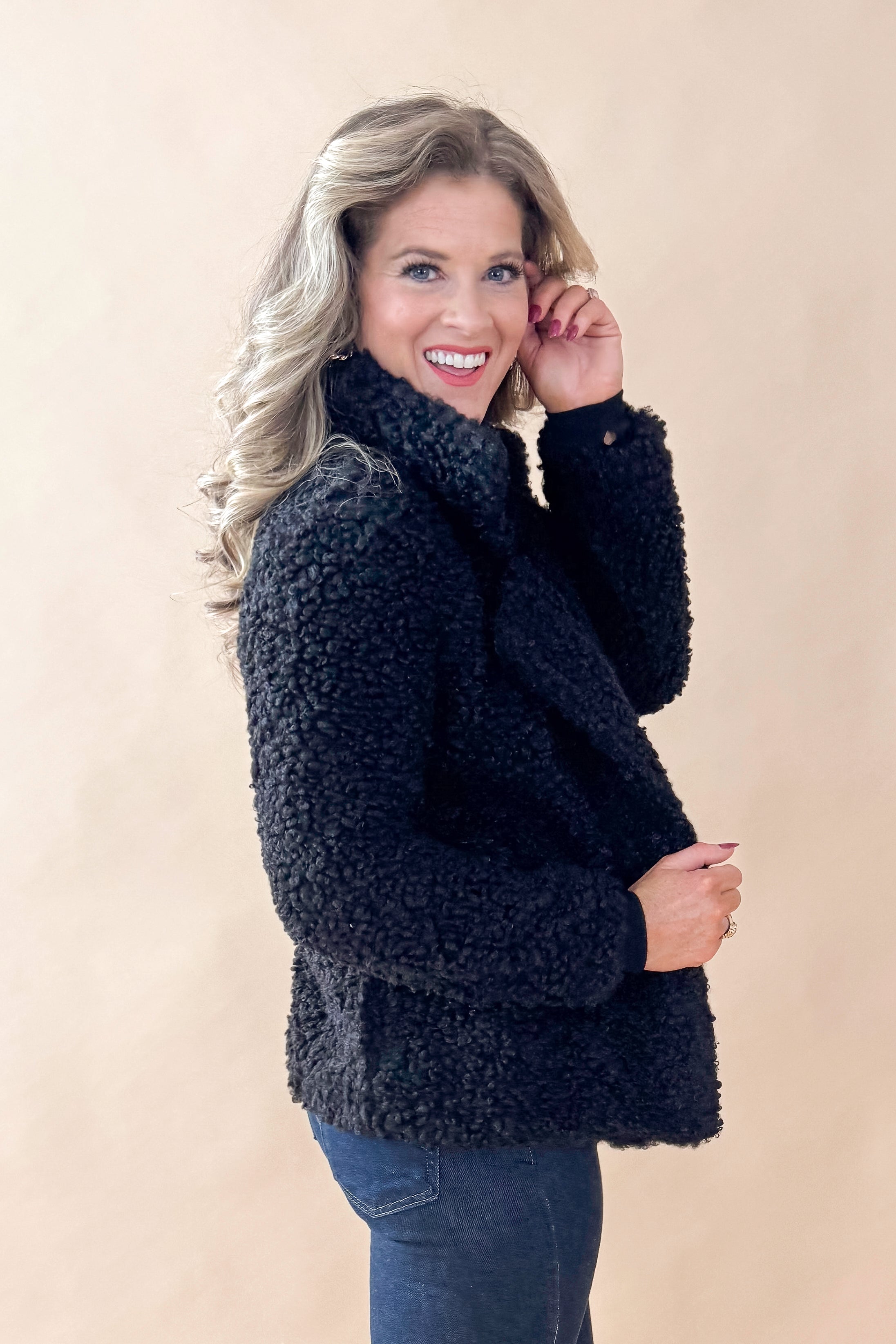 Side view of woman wearing Black Sherpa Coat and jeans against tan studio background.