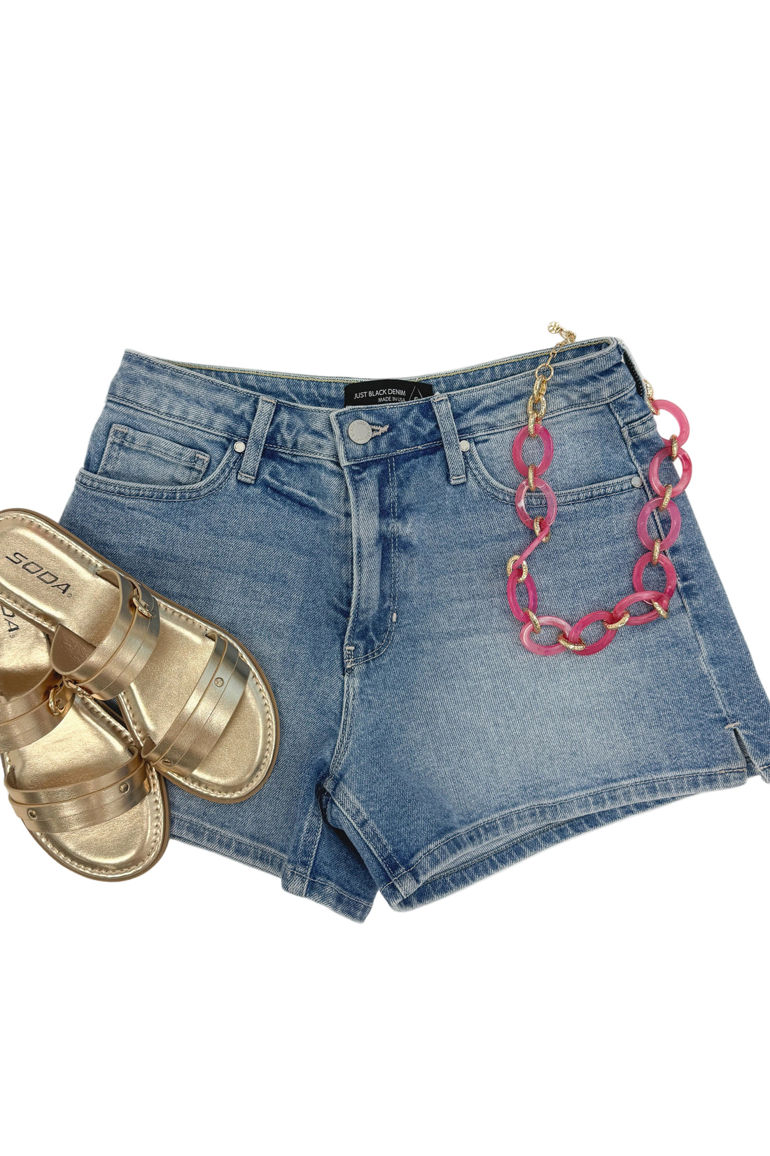 Flat lay of Swagger Boutique’s Light Wash Split Hem Denim Shorts by Just Black Denim with Maya Sandals and Indie Linked Necklace against white studio background.