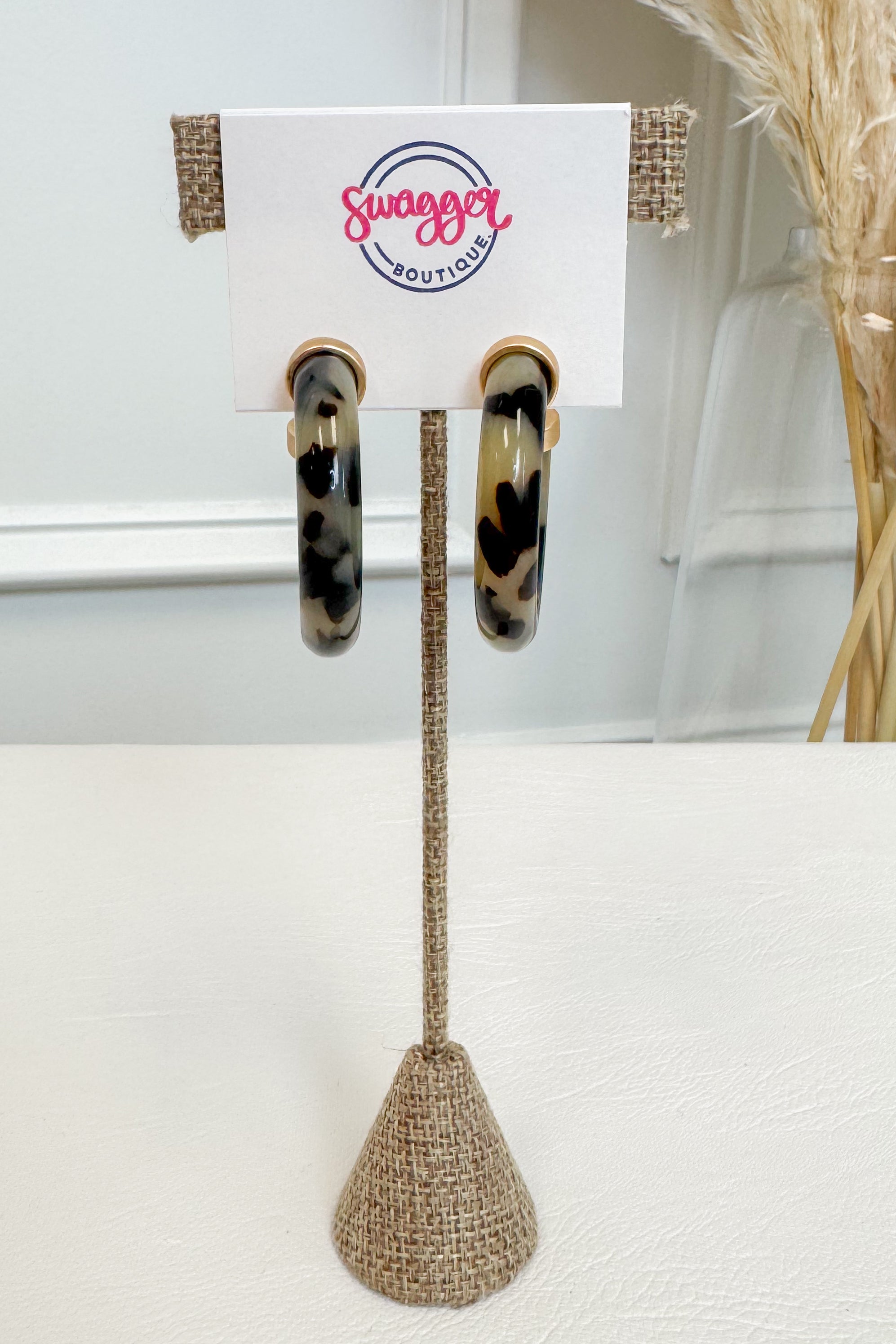 A pair of hoop earrings with a black, brown, and beige leopard print pattern, displayed on a white card stand with the &