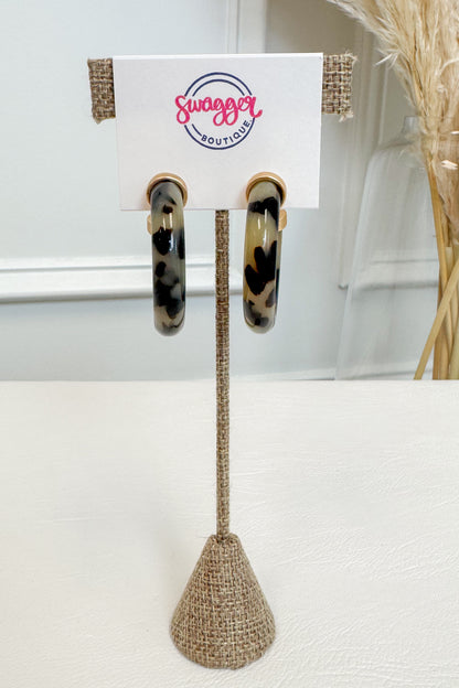 A pair of hoop earrings with a black, brown, and beige leopard print pattern, displayed on a white card stand with the &