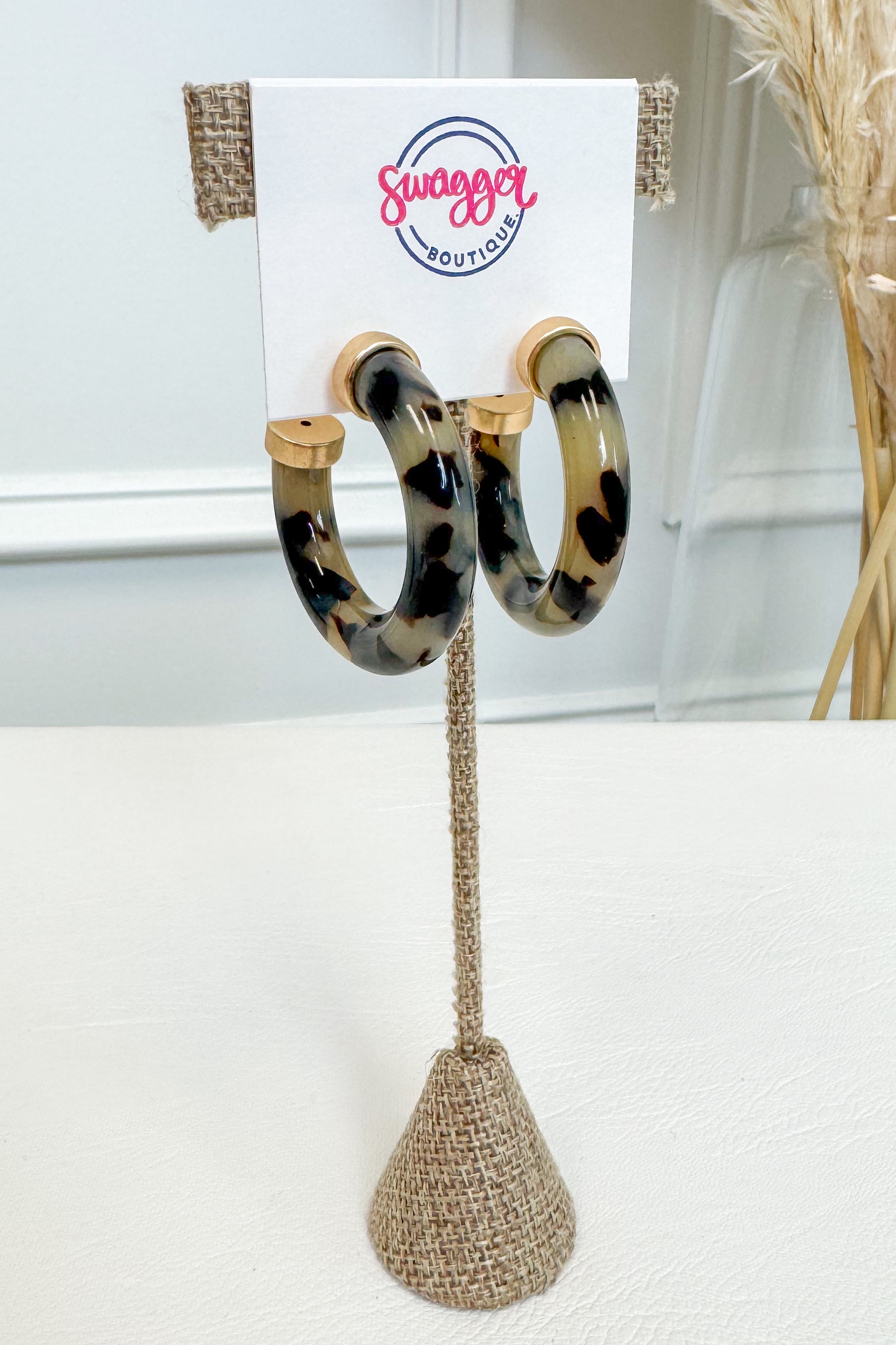 Tortoiseshell hoop earrings displayed on a white card stand with a burlap-wrapped stem, set against a softly blurred indoor background.