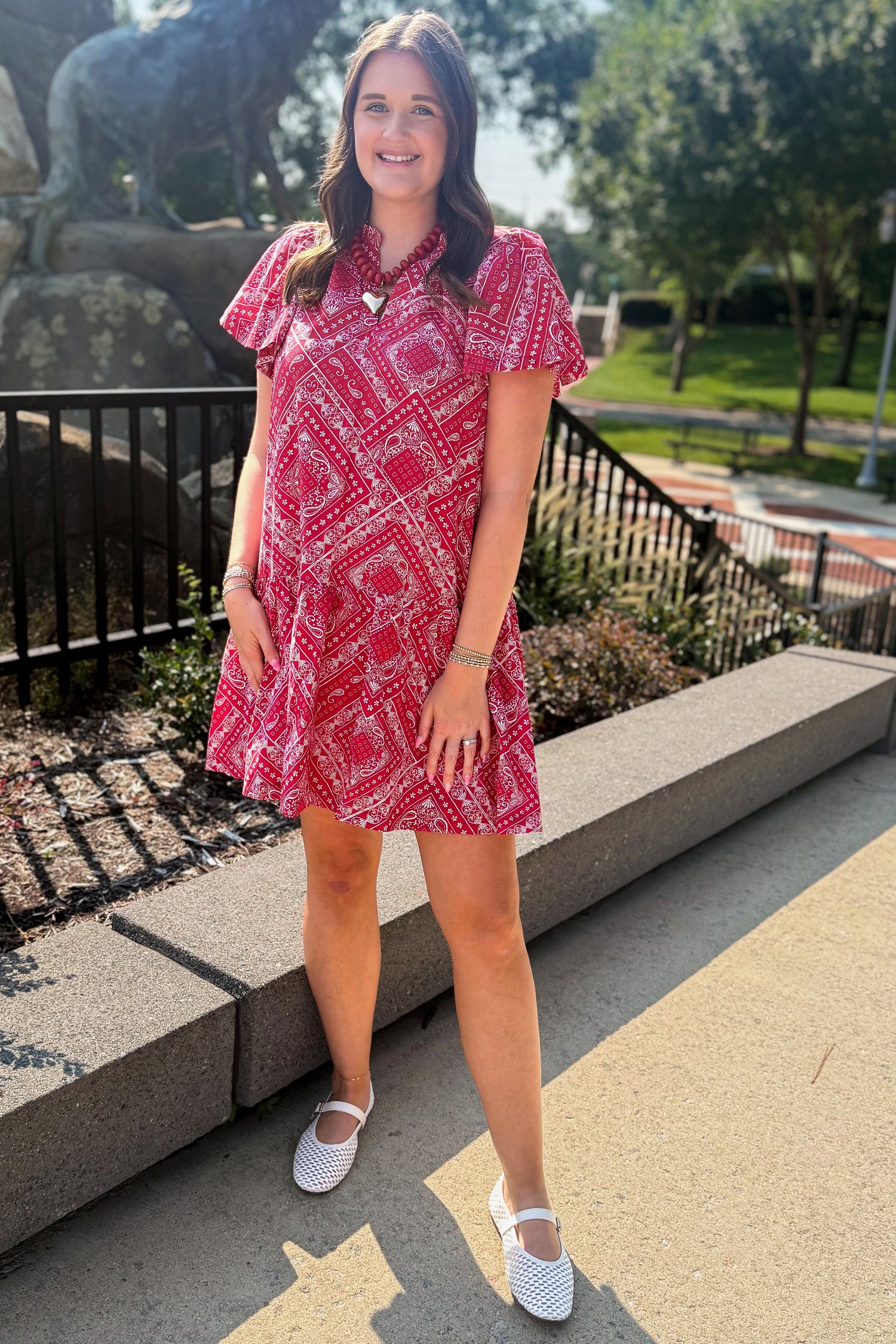 Front view of woman wearing Swagger Boutique’s Red Bandana Print Mini Dress standing outside at a park.