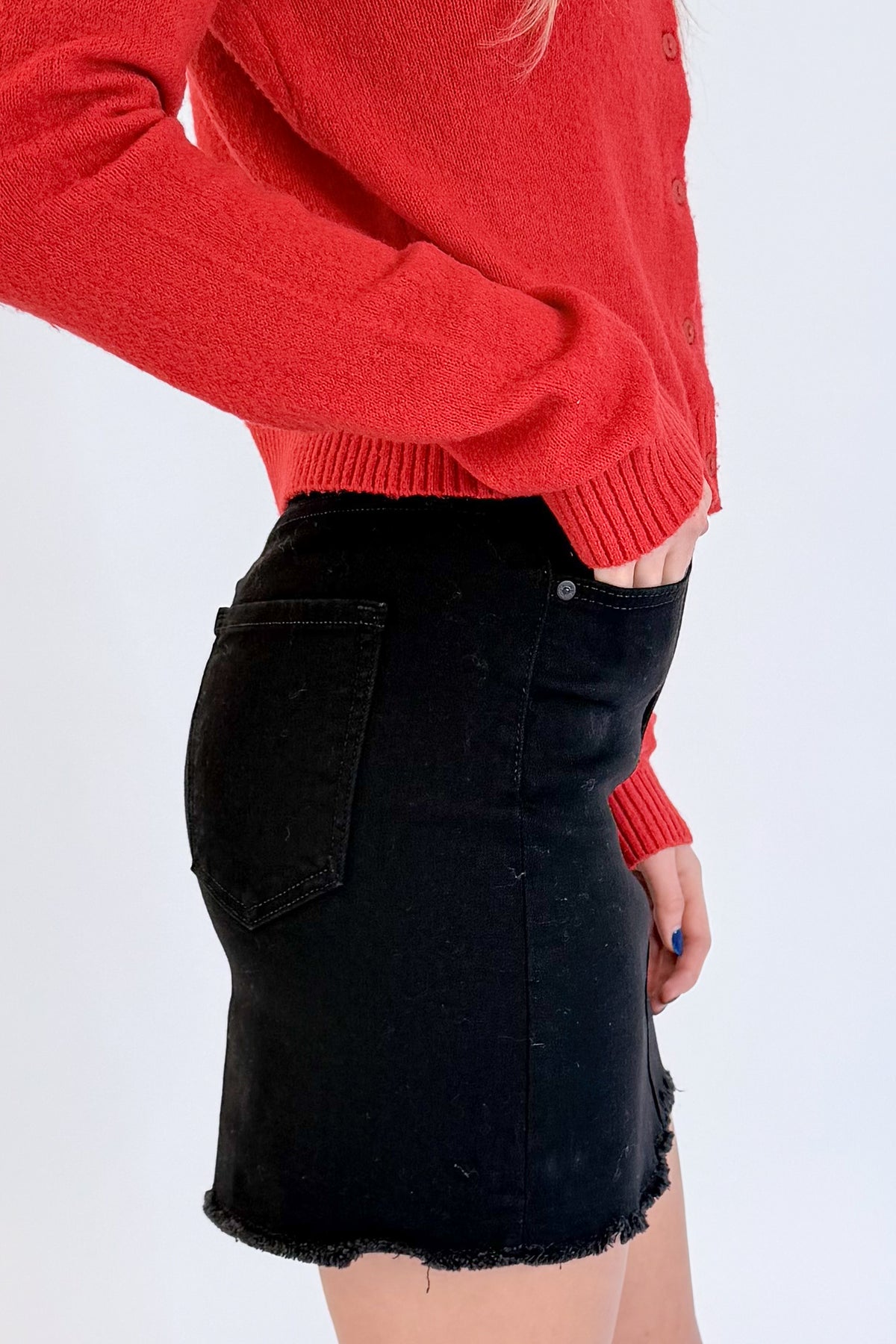 Side detail view of woman wearing Swagger Boutique’s Red Button Up Cardigan with Black Denim Mini Skirt by Just Black Denim against white studio background.