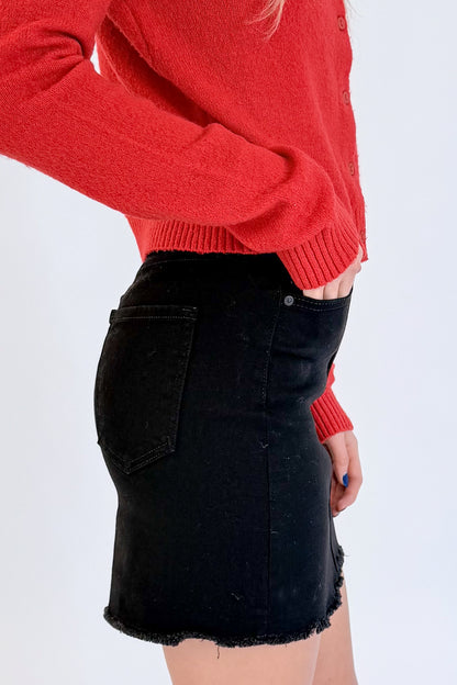 Side detail view of woman wearing Swagger Boutique’s Red Button Up Cardigan with Black Denim Mini Skirt by Just Black Denim against white studio background.