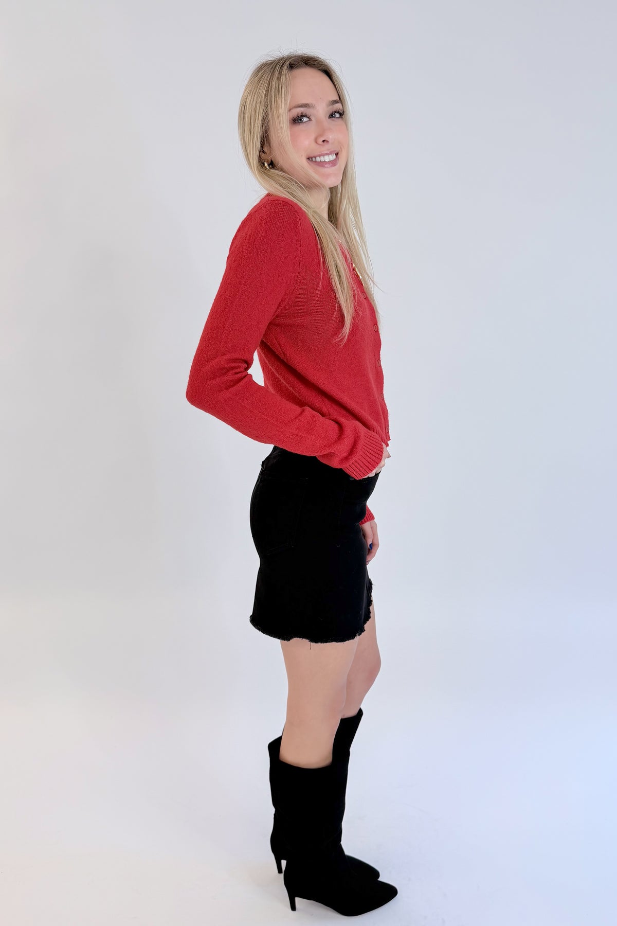 Side view of woman wearing Swagger Boutique’s Red Button Up Cardigan with Black Denim Mini Skirt by Just Black Denim against white studio background.