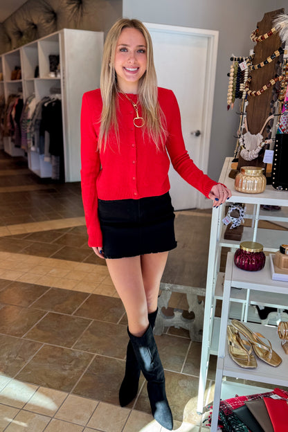 Front view of woman wearing Swagger Boutique’s Red Button Up Cardigan with Black Denim Mini Skirt by Just Black Denim standing at the store.