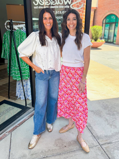 Front view of woman wearing Swagger Boutique’s Red Floral Print Tiered Midi Skirt with white sweater and woman wearing White Puff Sleeve Eyelet Top standing outside of Swagger boutique.