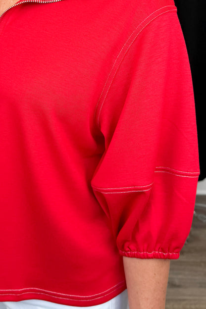 Front detail view of woman wearing Swagger Boutique’s Red Puff Sleeve 1_4 Zip Sweatshirt with jeans with clothing hanging in the background.