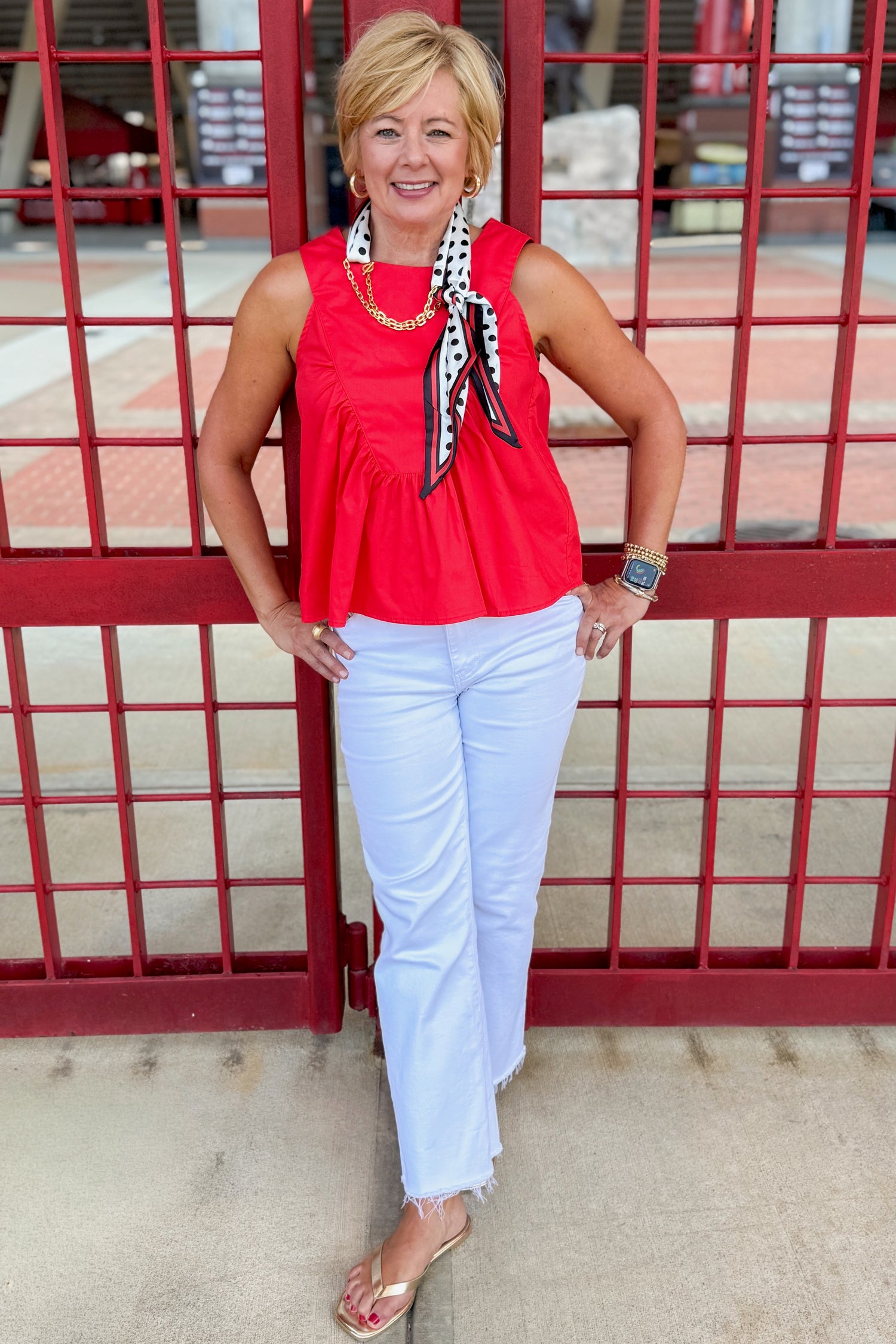 Front view of woman wearing Swagger Boutique’s Red Ruffle Tank with jeans standing by a stadium gate.