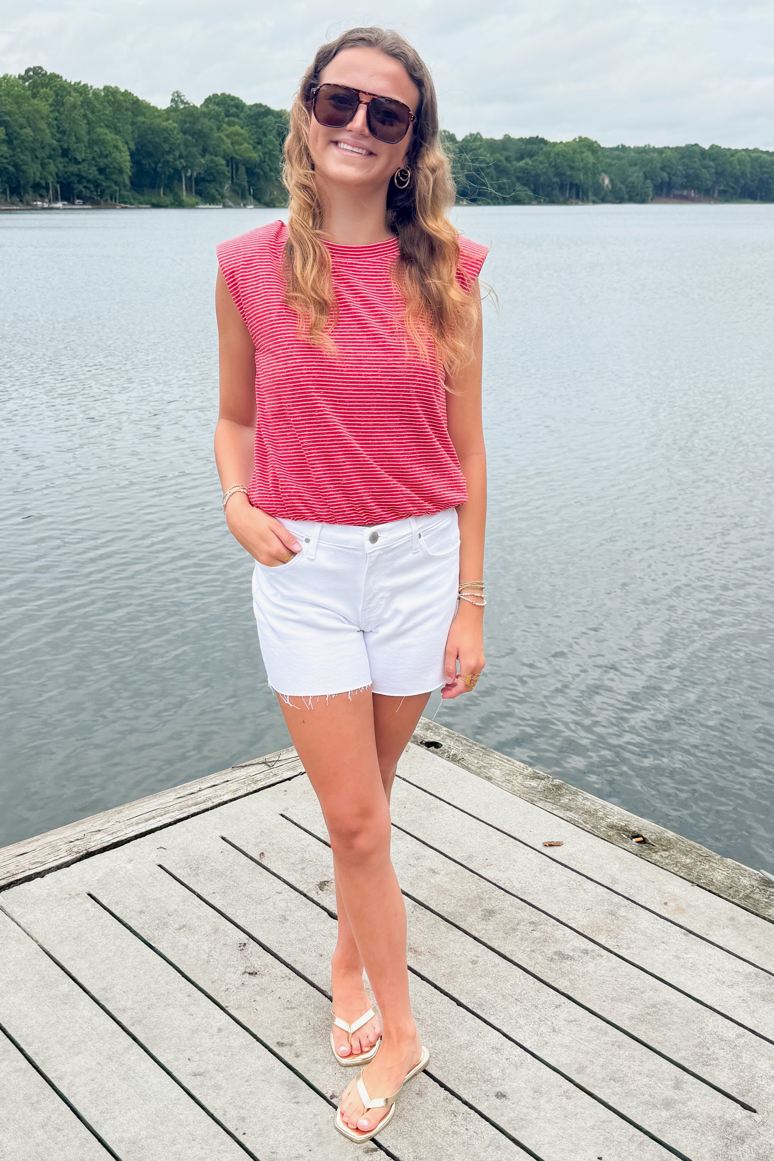 Front view of woman wearing Swagger Boutique’s Red Striped Bubble Hem Sleeveless Top with jean shorts standing on a dock at the lake.