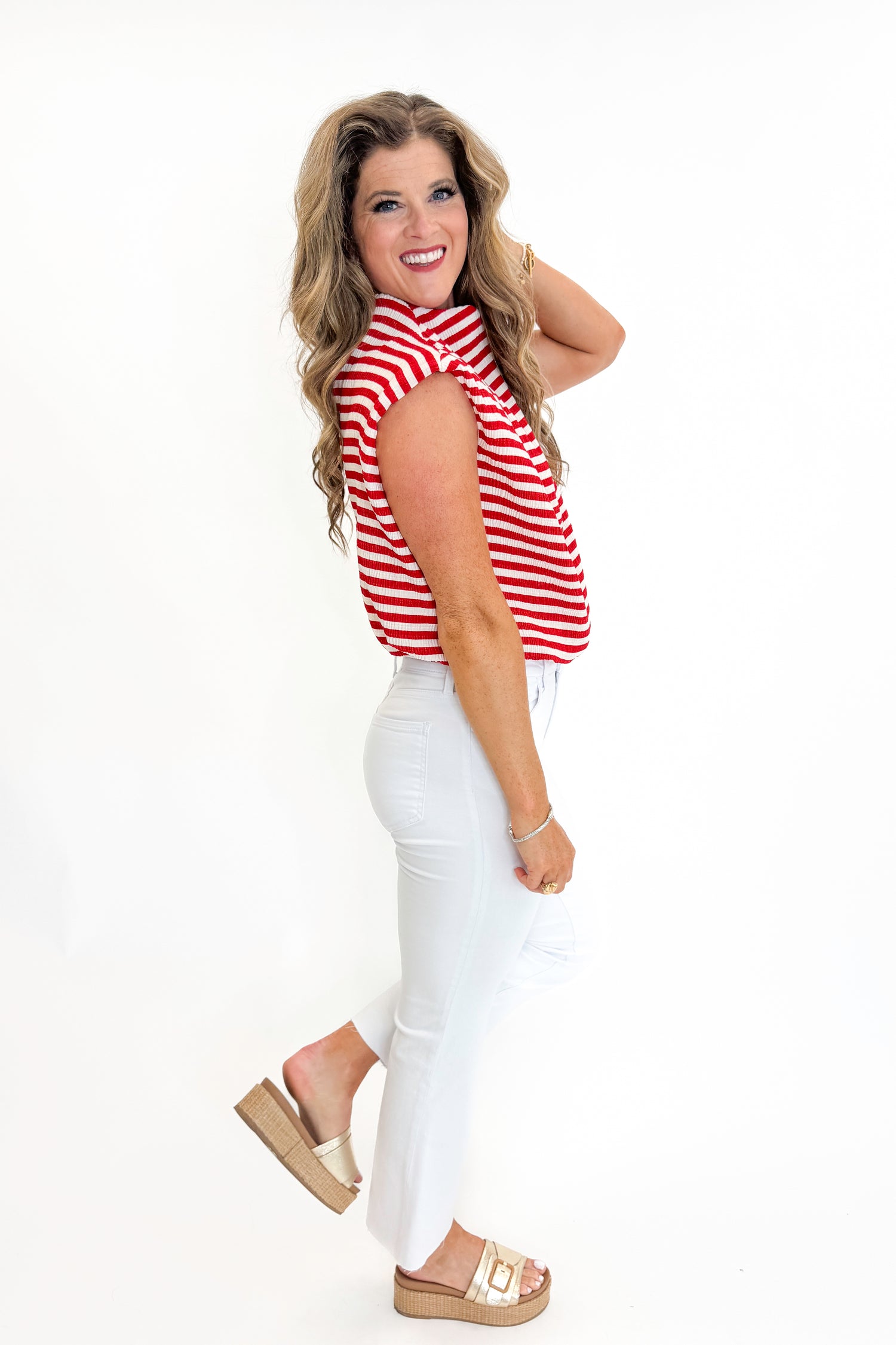 Side view of woman wearing Swagger Boutique’s Red Striped Sleeveless High Neck Top with jeans against white studio background.