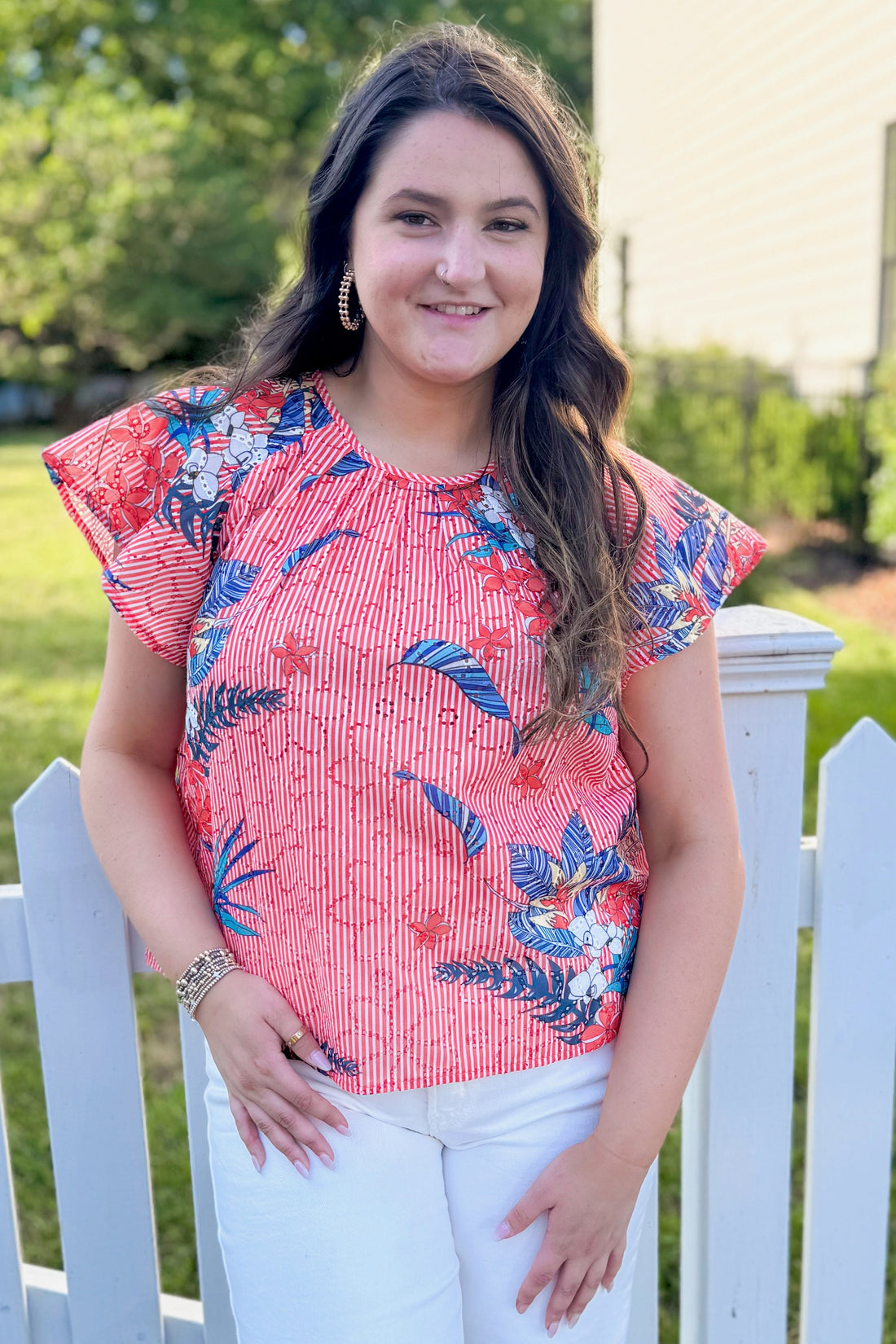 Front view of woman wearing Swagger Boutique’s Red Striped &amp; Floral Short Sleeve Top with jeans standing outside by a fence.