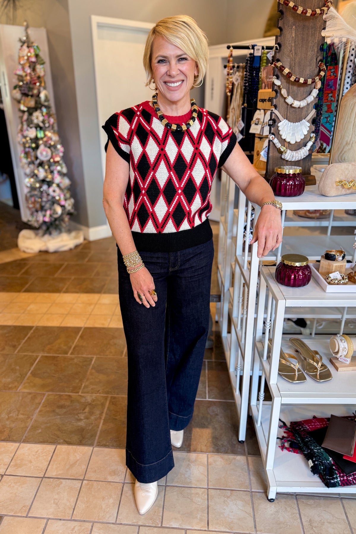 A smiling blonde woman stands at Swagger Boutique wearing a sleeveless knit top with a red, black, and white diamond argyle pattern, paired with black pants. 