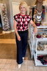 A smiling blonde woman stands at Swagger Boutique wearing a sleeveless knit top with a red, black, and white diamond argyle pattern, paired with black pants. 