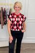 A smiling blonde woman stands indoors wearing a sleeveless knit top with a red, black, and white diamond argyle pattern, paired with black pants. A clothing rack with other garments is visible behind her against a white paneled wall.