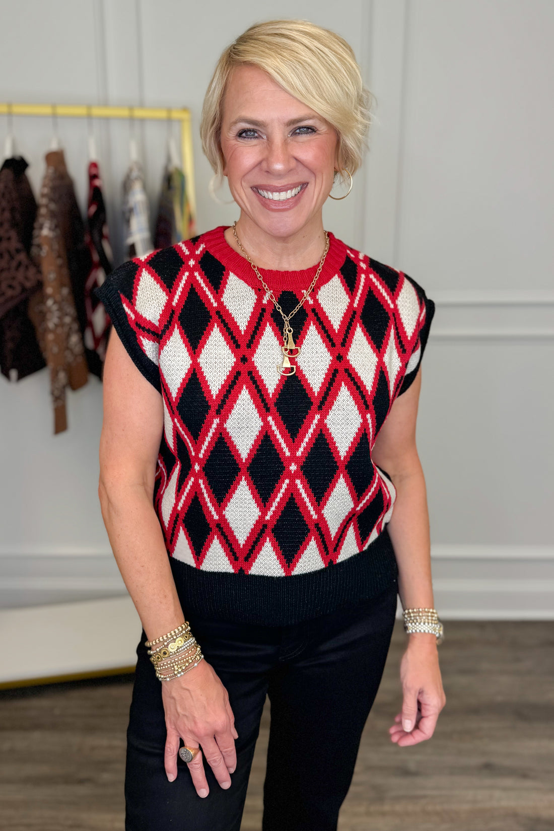 A smiling blonde woman wearing a red, black, and white argyle knit sweater vest over a red top and black pants, standing in a boutique setting.