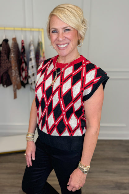 A smiling blonde woman wearing a red, black, and white argyle knit sweater vest with black pants, standing in a boutique setting with clothing racks visible in the background.