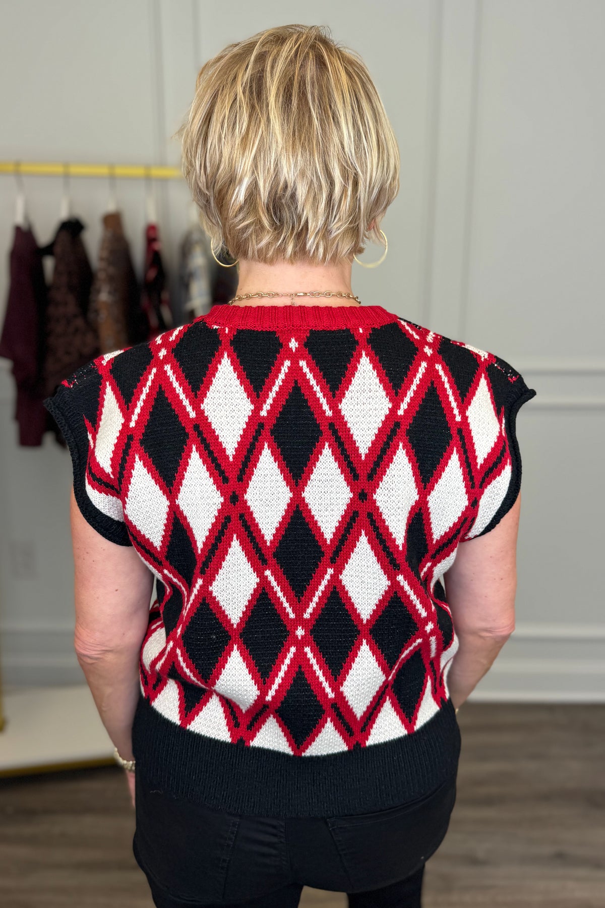 A woman viewed from behind wearing a short-sleeve knit sweater with a bold red, black, and white argyle diamond pattern. The sweater has a solid red crew neck and black sleeves.