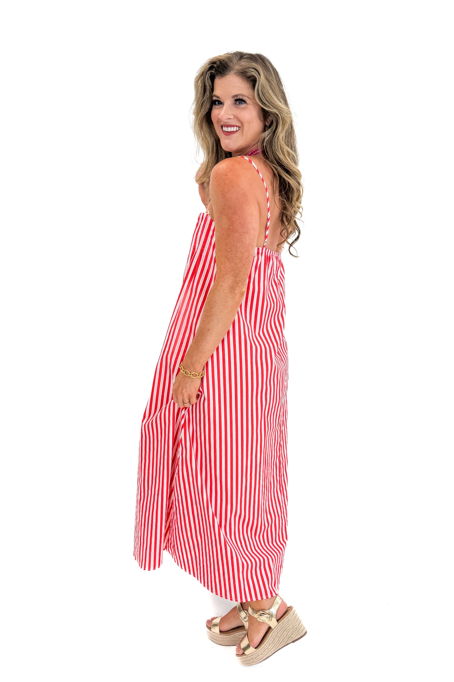 Side view of woman wearing Swagger Boutique’s Red &amp; Pink Striped Maxi Dress with sandals against white studio background.