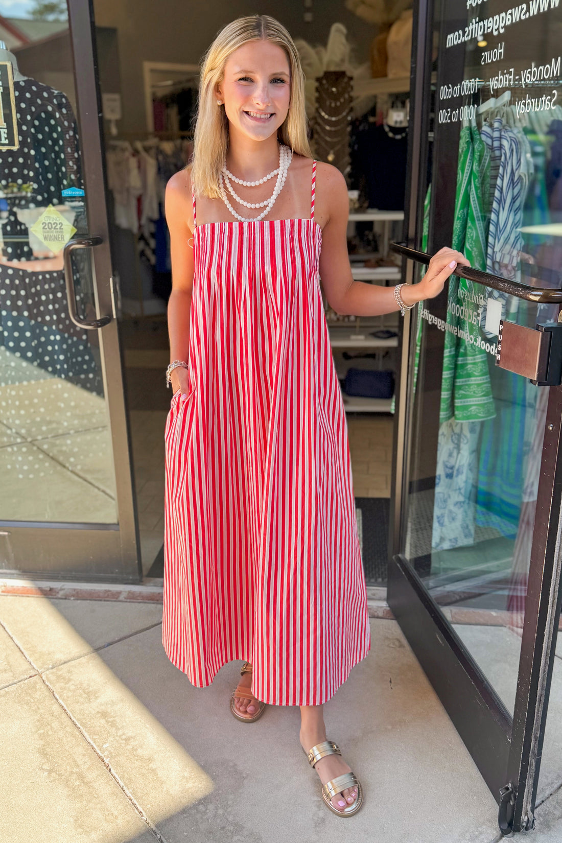 Front view of woman wearing Swagger Boutique’s Red &amp; Pink Striped Maxi Dress with sandals standing outside the store.