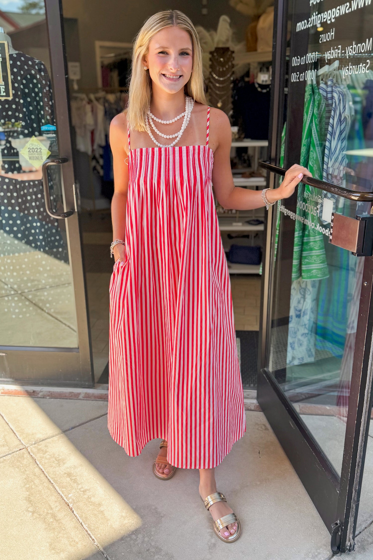 Front view of woman wearing Swagger Boutique’s Red &amp; Pink Striped Maxi Dress with sandals standing outside the store.