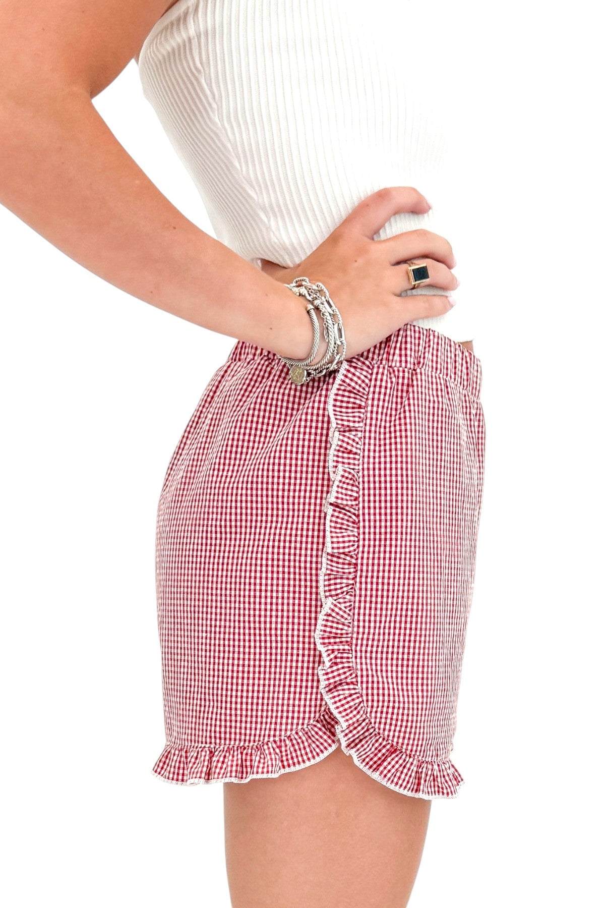 Side detail view of woman wearing Swagger Boutique’s Red &amp; White Gingham Ruffle Shorts with white top against white studio background.