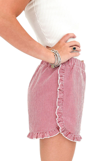 Side detail view of woman wearing Swagger Boutique’s Red &amp; White Gingham Ruffle Shorts with white top against white studio background.