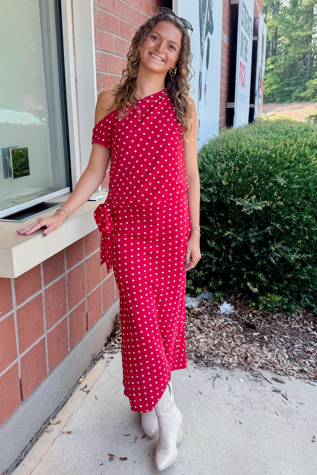 Front view of woman wearing Swagger Boutique’s Red &amp; White Polka Dot Midi Skirt with Red &amp; White Polka Dot Asymmetrical Tank standing outside a ticket office.
