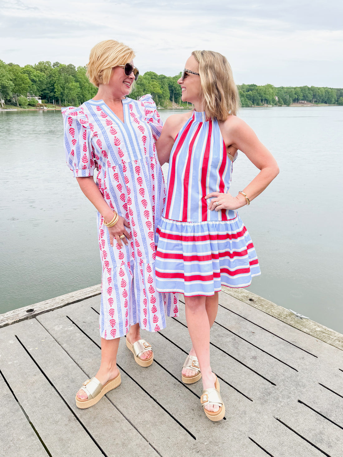 Front view of woman wearing Swagger Boutique’s Red, White &amp; Blue Jonah Midi Dress by MudPie with Lulah Platform Sandals and sunglasses with woman wearing Blue &amp; Red Striped Halter Neck Dress by Entro outside at the lake.