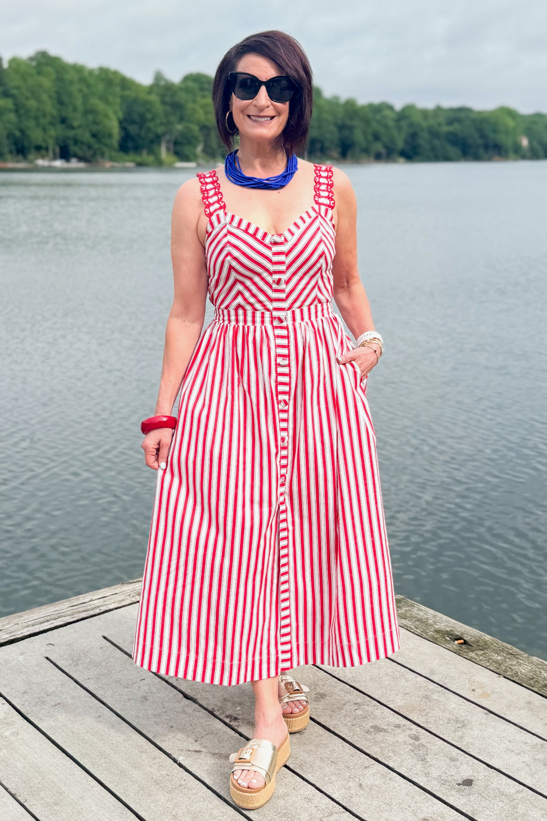 Front view of woman wearing Swagger Boutique’s Red, White &amp; Blue Striped Midi Dress with sandals standing outside at the lake.