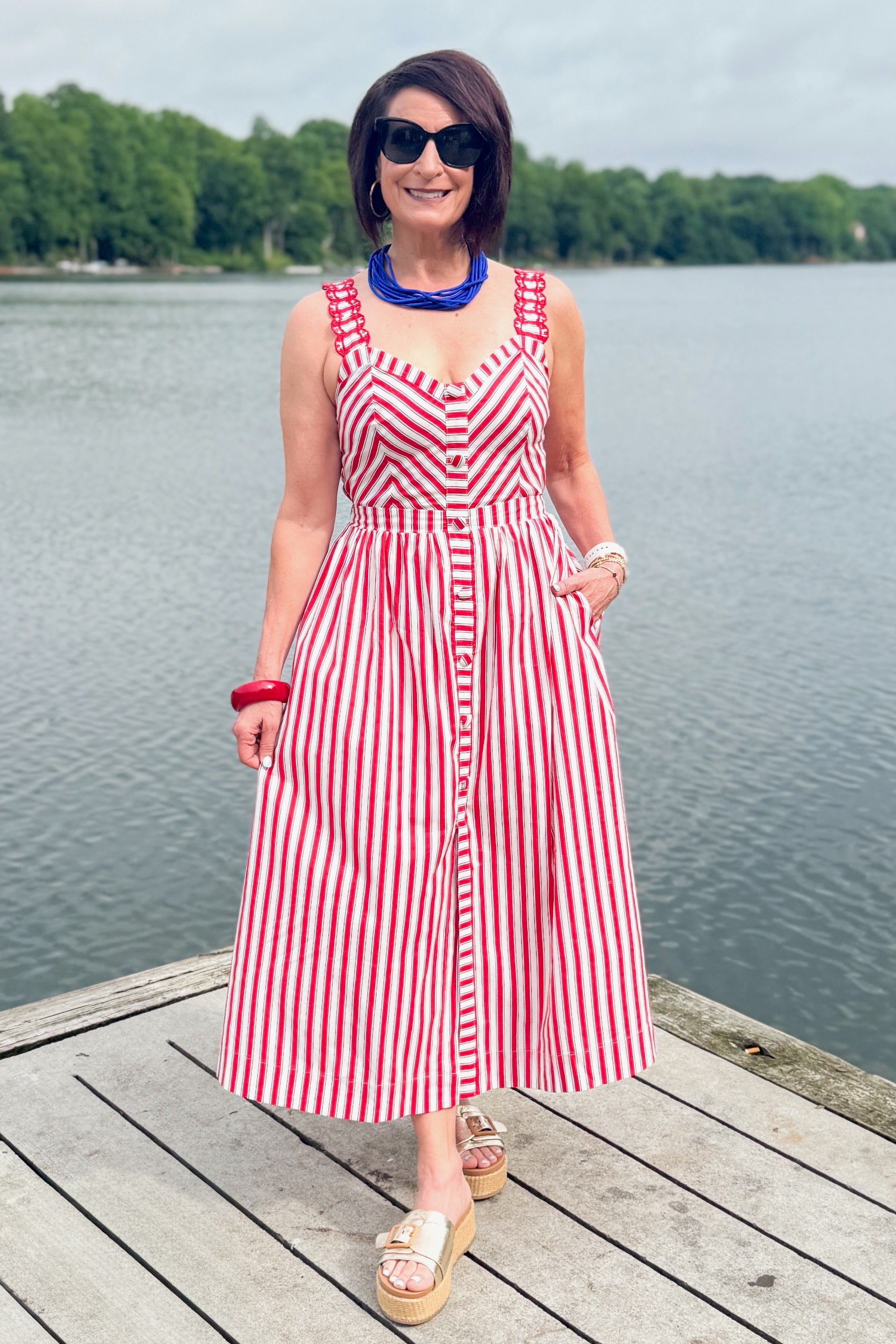 Front view of woman wearing Swagger Boutique’s Red, White &amp; Blue Striped Midi Dress with sandals standing outside at the lake.