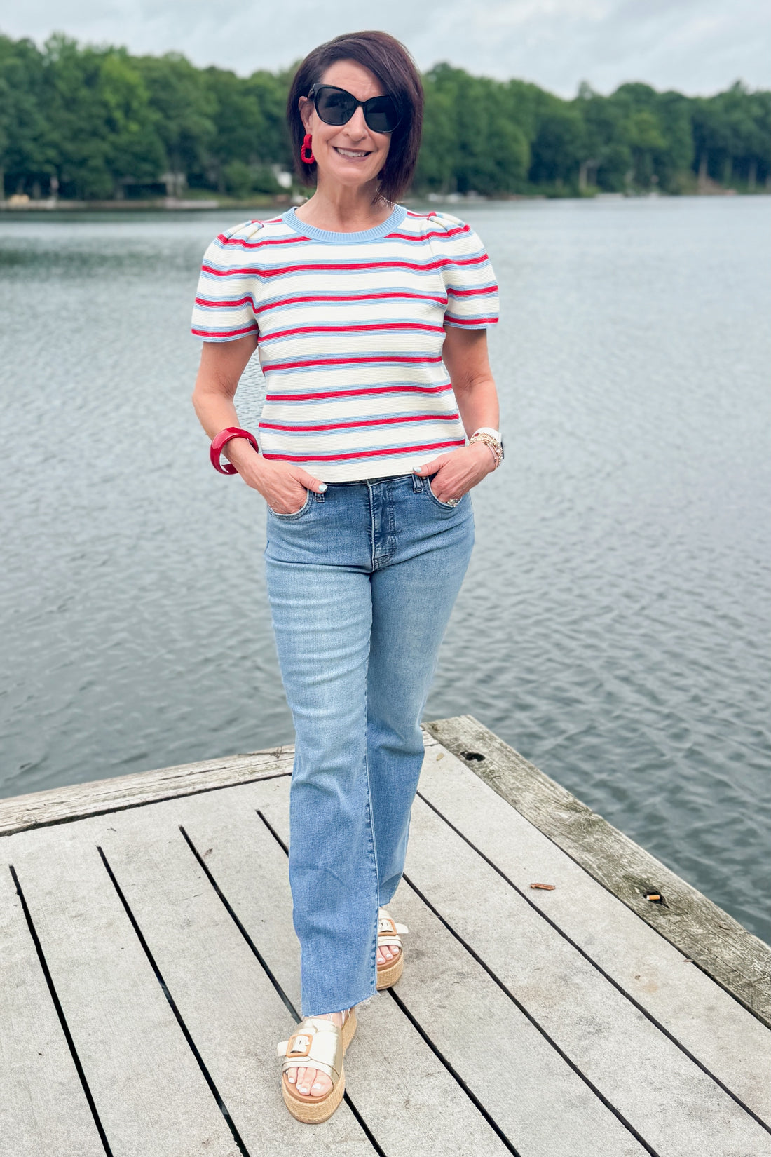 Front view of woman wearing Swagger Boutique’s Red, White &amp; Blue Striped Top by Entro with jeans standing on a dock outside.
