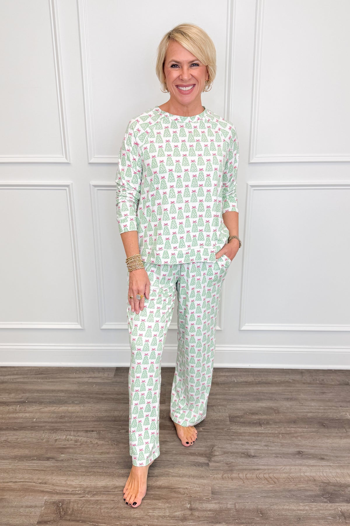 Model wearing the Rosy Woodlands Annie Pajama Set, featuring an allover green Christmas tree print with pink bows on a white background. The set includes a relaxed-fit, long-sleeve crewneck top and matching straight-leg pajama pants with side pockets.