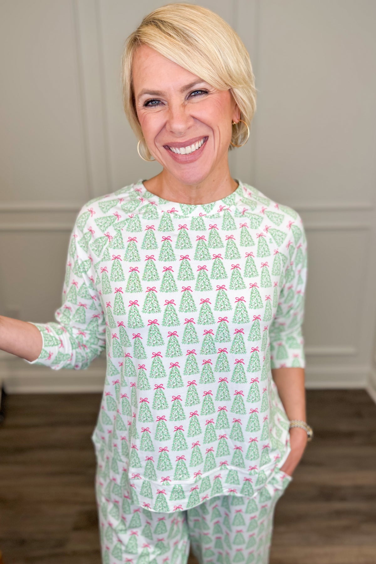 Model wearing the Rosy Woodlands Annie Pajama Set, featuring an allover green Christmas tree print with pink bows on a white background. The set includes a relaxed-fit, long-sleeve crewneck top and matching straight-leg pajama pants with side pockets.