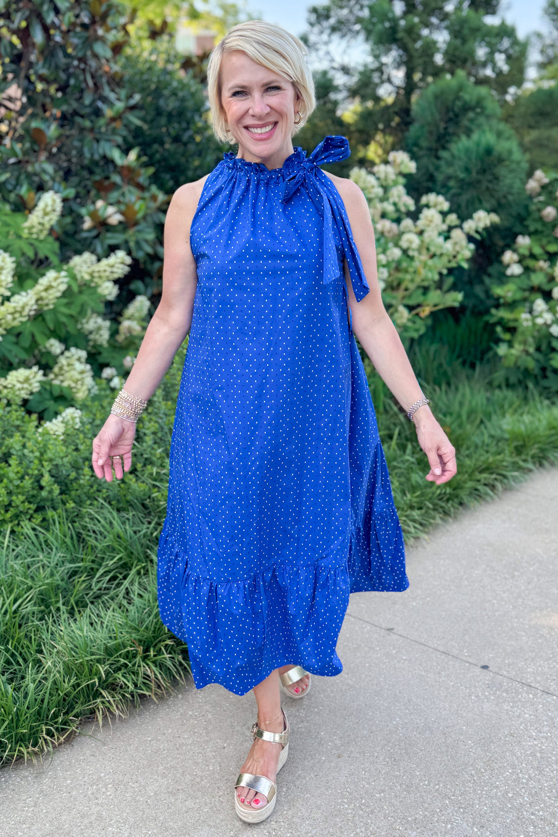 Front view of woman wearing Swagger Boutique’s Royal Blue Polka Dot Tue Neck Midi Dress walking outside on a path.