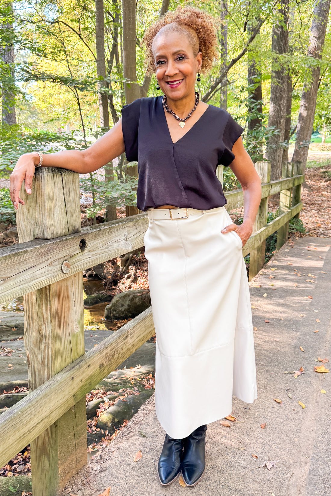Front view of woman outside wearing Ecru Faux Leather Midi Skirt and black top with trees in the background.