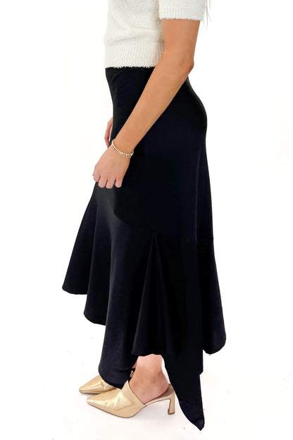 Side view of woman wearing Black Satin Handkerchief Hem Midi Skirt with sweater against white studio background.