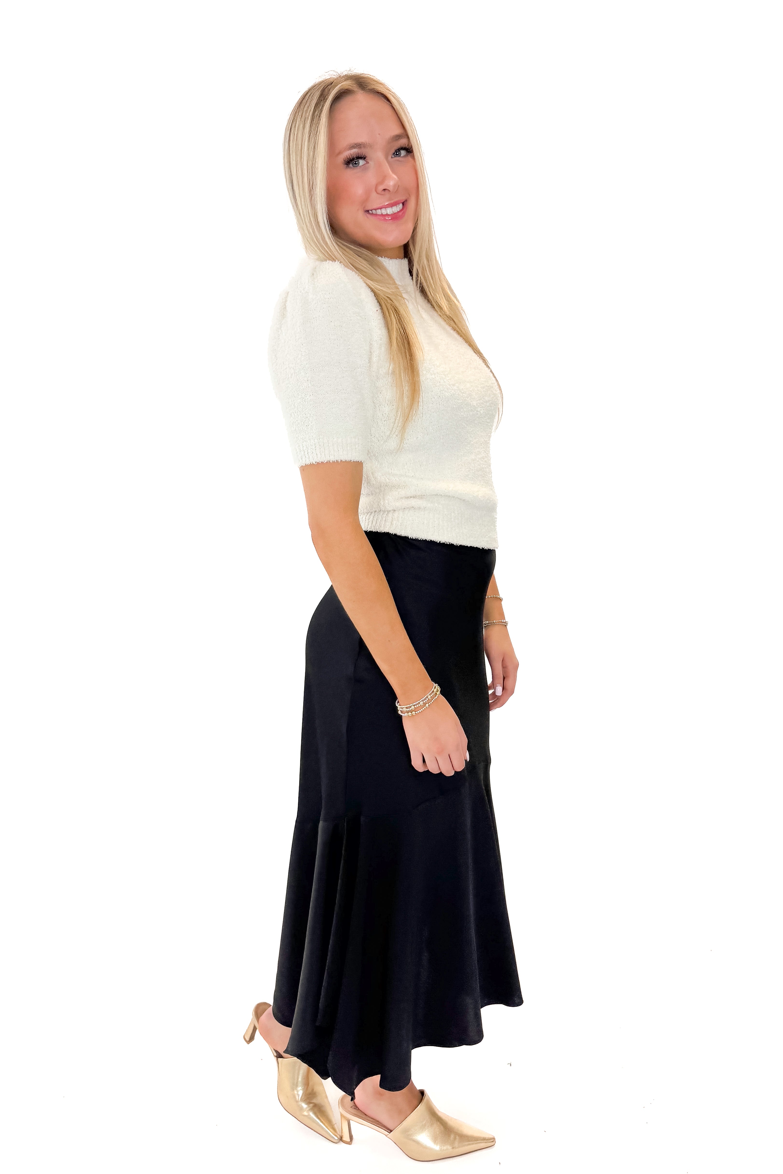 Side view of woman wearing Black Satin Handkerchief Hem Midi Skirt with sweater against white studio background.
