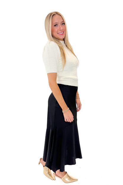 Side view of woman wearing Black Satin Handkerchief Hem Midi Skirt with sweater against white studio background.