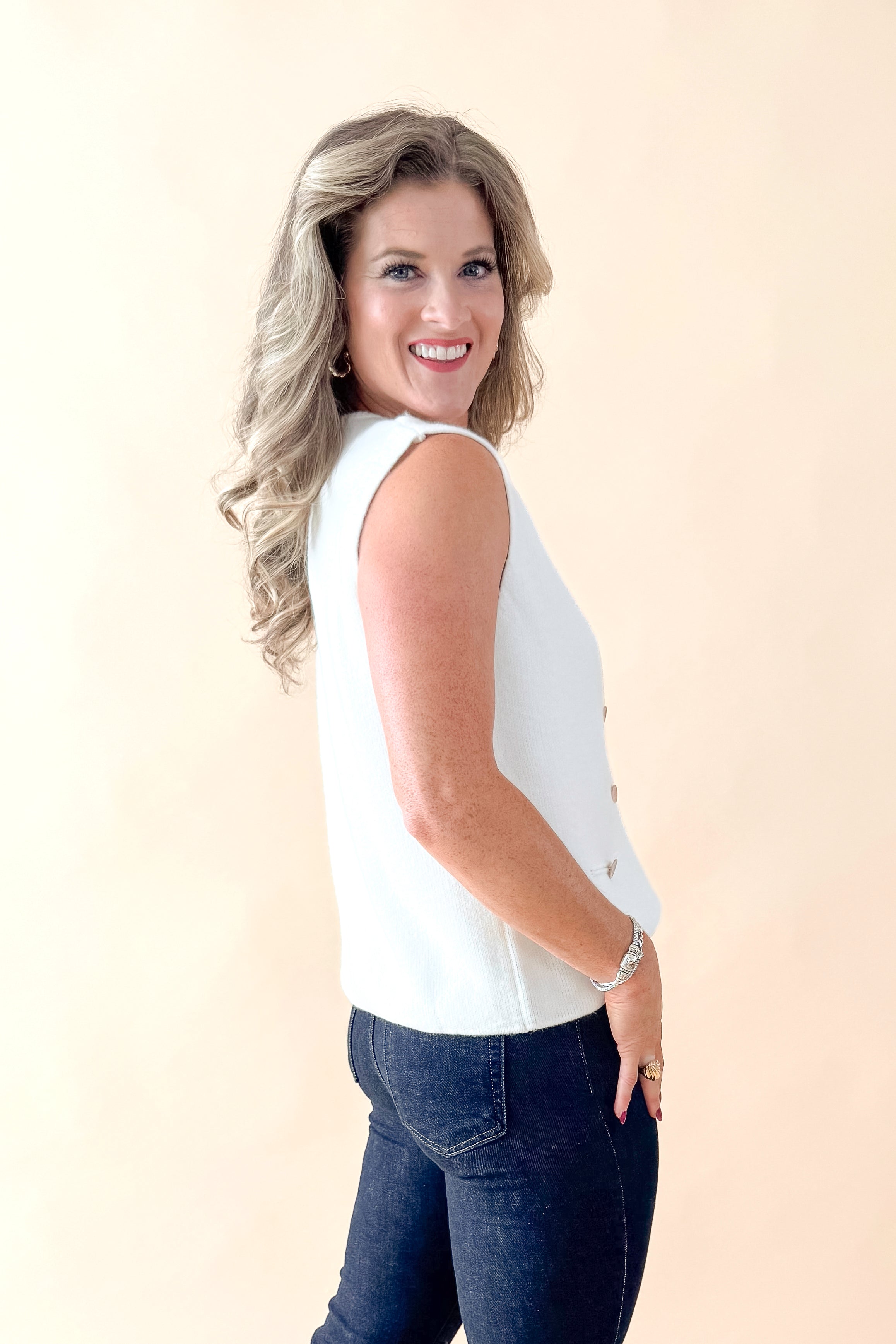 Side view of woman wearing Cream Button Detail Sweater Vest with jeans against tan studio background.